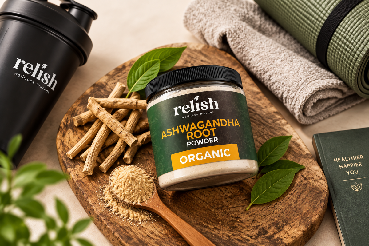 A jar of Relish Market Organic Ashwagandha Root Powder sits on a wooden cutting board with root pieces, a wooden spoon of powder, green leaves, a black Relish shaker bottle, a folded towel, and a book in a styled tabletop scene.