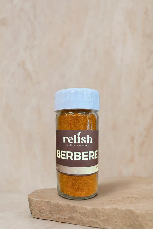 A small clear plastic bottle of Relish Market Berbere seasoning with a white cap and a brown label, sitting on a textured beige stone slab against a matching beige background.