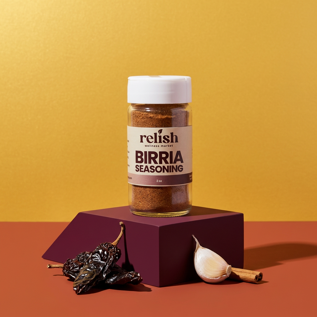 A bottle of Relish Market Birria Seasoning stands on a dark purple geometric pedestal against a warm yellow background, with dried chilies and a garlic bulb placed at the base.