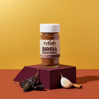 A bottle of Relish Market Birria Seasoning stands on a dark purple geometric pedestal against a warm yellow background, with dried chilies and a garlic bulb placed at the base.