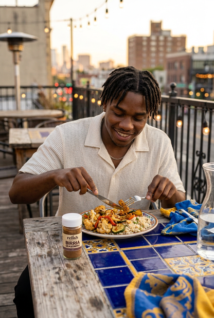 A man wearing a cream ribbed short-sleeve button-up shirt sits at a wooden table on a rooftop patio, cutting into a plate of food. A bottle of Birria Seasoning by Relish Market is visible on the table next to a blue and gold patterned napkin, with a city skyline in the background at sunset.