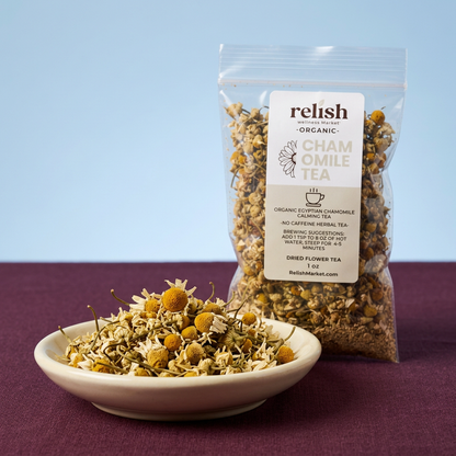 A clear stand-up pouch of Relish Market Organic Chamomile Tea stands behind a small white bowl filled with loose dried chamomile flowers on a dark purple surface against a light blue background.