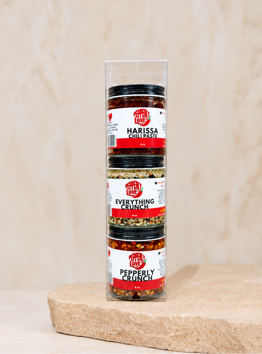 A clear cylindrical spice jar package containing three stacked jars of Harissa Chili Paste, Everything Crunch, and Peppery Crunch, displayed on a stone slab against a neutral beige background.