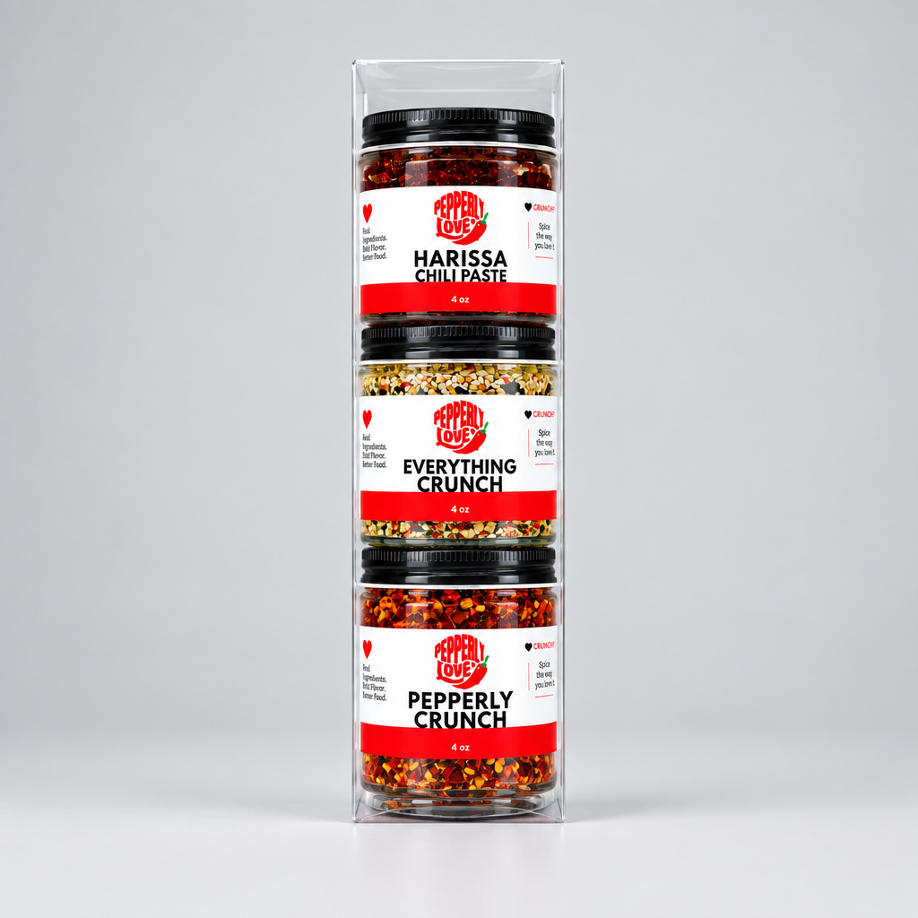 A three-pack of condiment jars in a clear plastic display box, set against a neutral light gray background. The jars contain Harissa Chili Paste, Everything Crunch, and Pepperly Crunch chili  with black lids and white labels featuring red and black text.