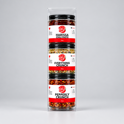 A three-pack of condiment jars in a clear plastic display box, set against a neutral light gray background. The jars contain Harissa Chili Paste, Everything Crunch, and Pepperly Crunch chili  with black lids and white labels featuring red and black text.