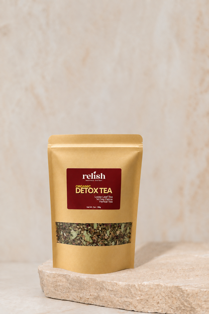 A stand-up pouch of Relish Market Organic Detox Tea placed on a stone slab against a textured beige background. The pouch is gold with a dark red label and a clear window showing the loose tea leaves inside.
