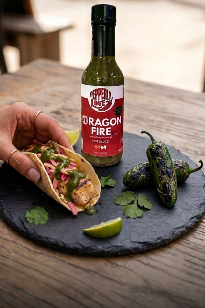 A bottle of Pepperly Love Dragon Fire hot sauce stands on a wooden table beside a hand holding a taco with green sauce, lime wedges, jalapeños, and cilantro on a dark slate platter.