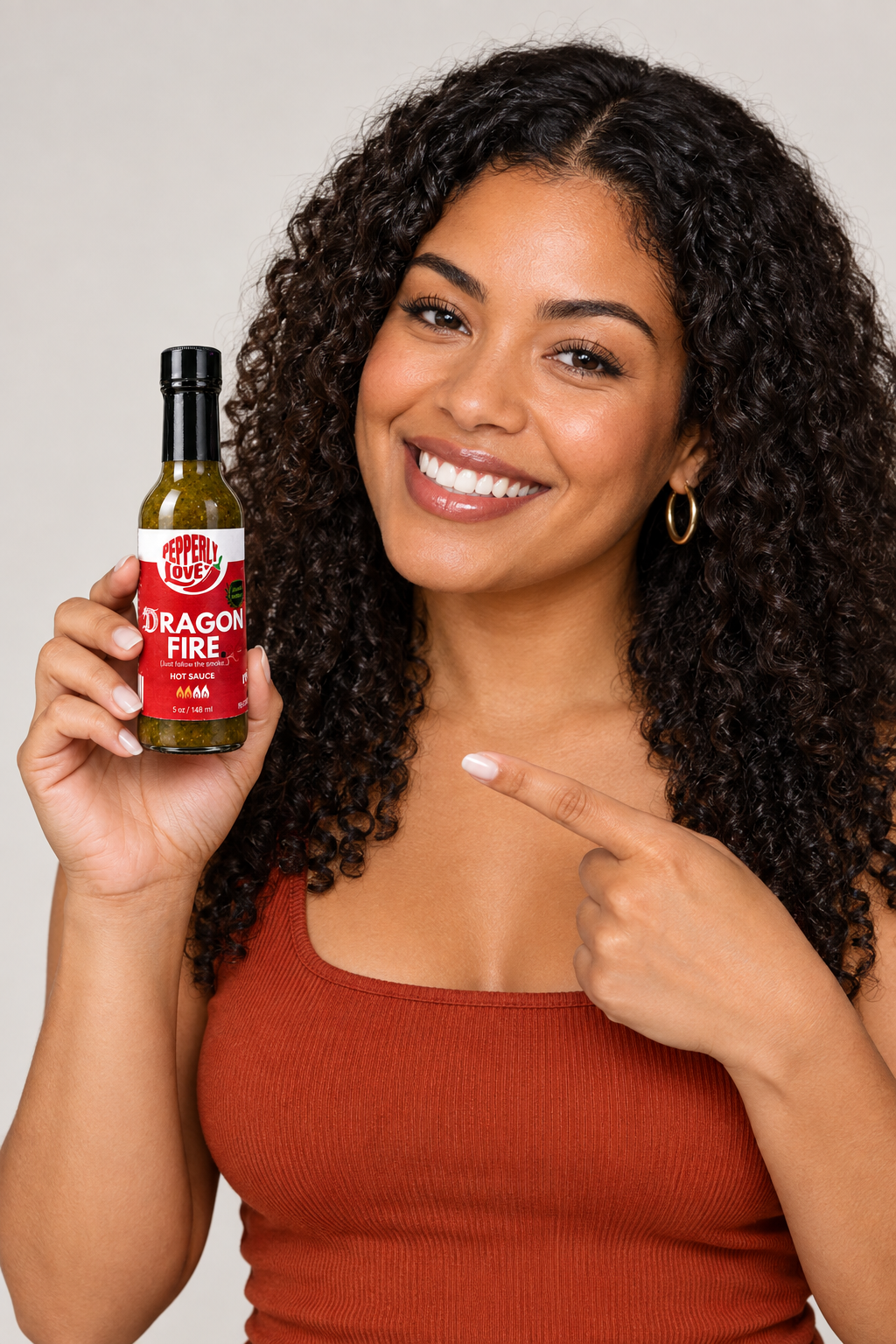 A smiling woman with curly dark hair holds up a bottle of Dragon Fire hot sauce, pointing at the label with her other hand, against a plain light gray background.