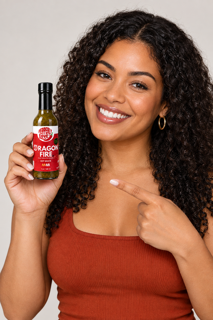 A smiling woman with curly dark hair holds up a bottle of Dragon Fire hot sauce, pointing at the label with her other hand, against a plain light gray background.