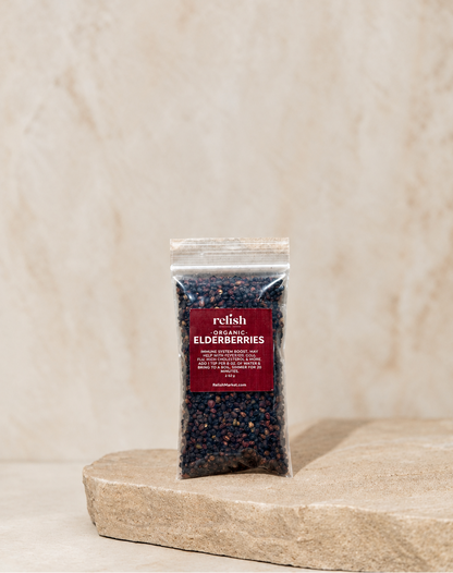A clear stand-up pouch containing dark blue-black elderberries, with a dark red label reading 'Organic ELDERBERRIES,' placed on a light stone slab against a soft beige textured background.
