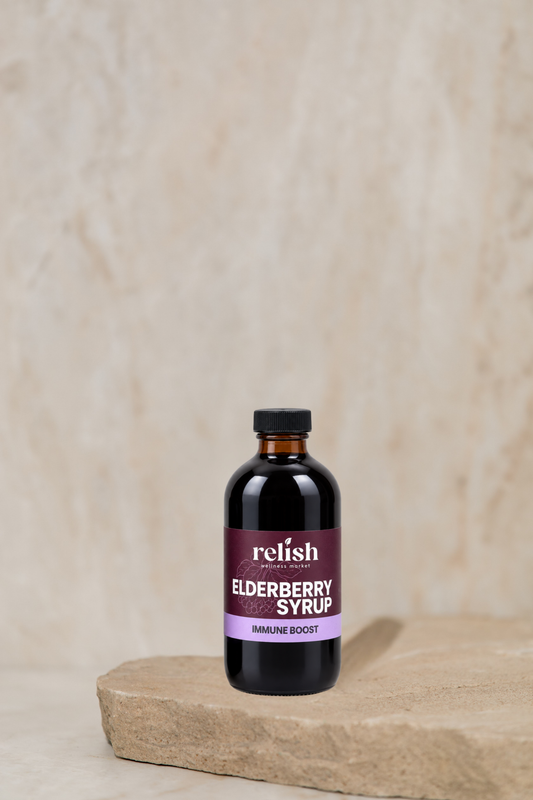 An amber colored bottle of Relish Market's Elderberry Syrup with an 'Immune Boost' label, placed on a rustic stone slab against a textured beige background.