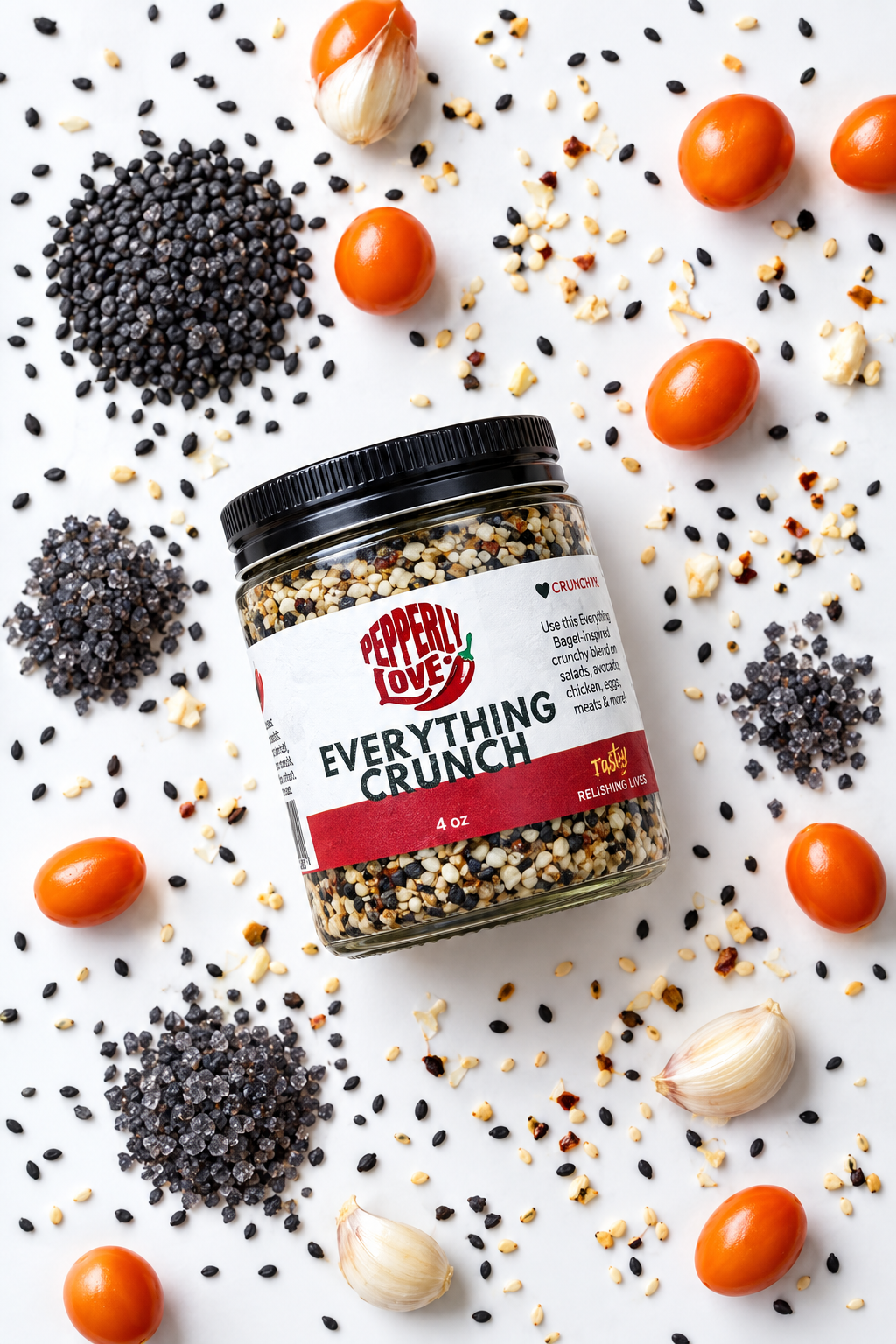 A 4 oz glass jar of Pepperly Love's Everything Crunch mix with a black lid and white label, set on a wood white background with black and white sesame seeds, red chili flakes, garlic cloves and red cherry tomatoes.