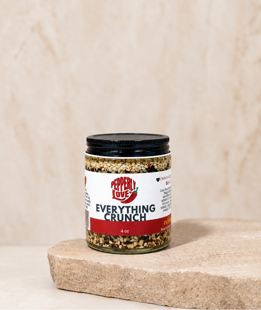 A small glass jar of Everything Crunch seasoning mix sits on a rustic stone slab against a textured beige background.