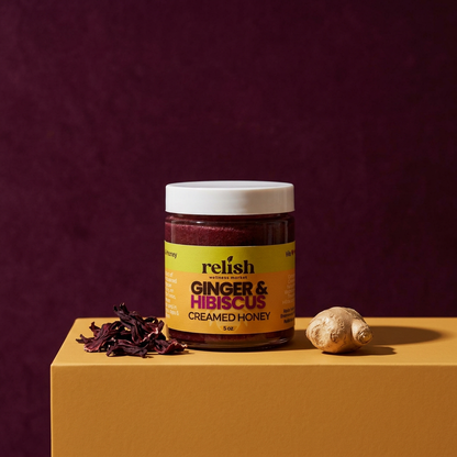 A jar of Ginger & Hibiscus Creamed Honey by Relish Market on a bold yellow surface, a bold plum background with pieces of hibiscus flowers and fresh ginger piece. Raw, unfiltered honey.
