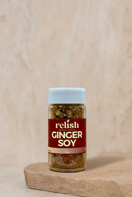 A bottle of Relish Market Ginger Soy seasoning standing on a stone slab against a textured beige background. The bottle has a white cap and a dark red label with white text.