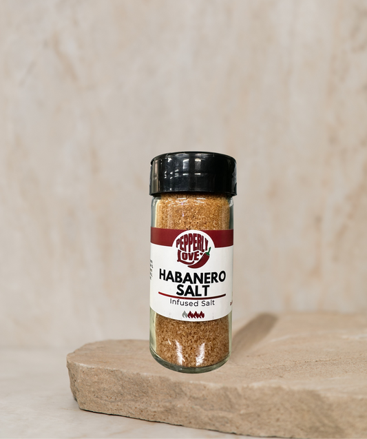 A bottle of Pepperly Love's Habanero Salt stands on a stone slab against a textured beige background. The clear glass bottle shows the brownish-orange salt inside, with a black lid and a white label featuring red and black text.