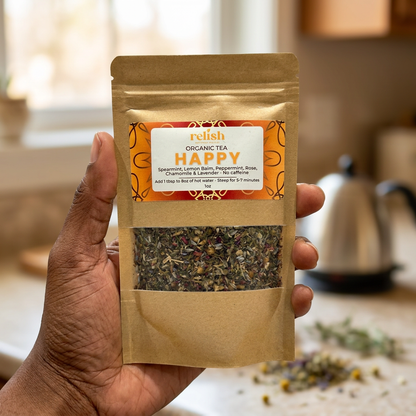 Black person's hand holding a bag of Relish Market Happy Organic Loose Leaf Tea in a kitchen with a stainless steel tea pot blurred in the background.