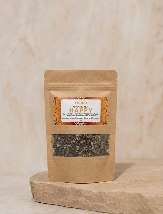 A Kraft stand-up pouch of Relish Market Happy Organic Tea placed on a light stone slab against a soft beige textured background. The pouch has a clear window showing loose-leaf tea inside and a front label reading 'HAPPY'.