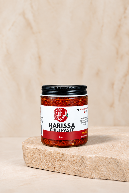 A jar of Pepper Love Harissa Chili Paste sits on a rustic stone slab against a textured beige background.