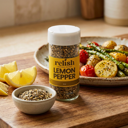 A jar of Relish Market'sLemon Pepper Seasoning close-up with plate of vegetables and lemon wedge.