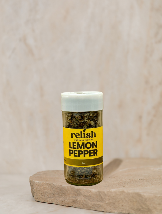 A bottle of Relish Market Lemon Pepper seasoning stands on a stone slab against a textured beige background. The clear bottle shows the greenish-yellow dried pepper flakes inside, with a bright yellow label and a white cap.