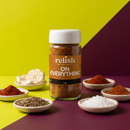 A jar of On Everything Spice Blend by Relish Market on a chartreuse and plum background, with the individual spice blend ingredients in small bowls placed around the jar.