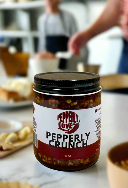 A jar of Pepperly Crunch by Pepperly Love sits on a kitchen counter top with 2 people cooking blurred in the background.