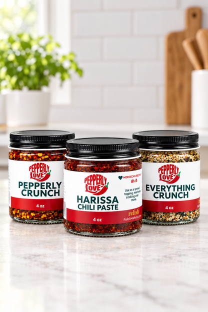 Three jars of Pepperly Love hot sauces arranged on a marble countertop in a bright kitchen setting. The center jar is Harissa Chili Paste, flanked by Pepperly Love Peppery Crunch and Pepperly Love Everything Crunch jars.