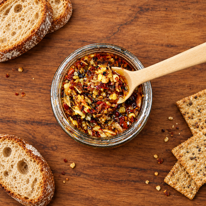 A small glass jar of Pepperly Love's Pepperly Crunch sits on a wooden table, with a wooden spoon resting inside. Slices of rustic bread are arranged around it on a warm wood surface.