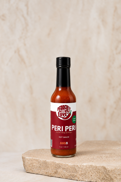 A bottle of Pepperly Love Peri Peri hot sauce stands on a light stone slab against a textured beige background.