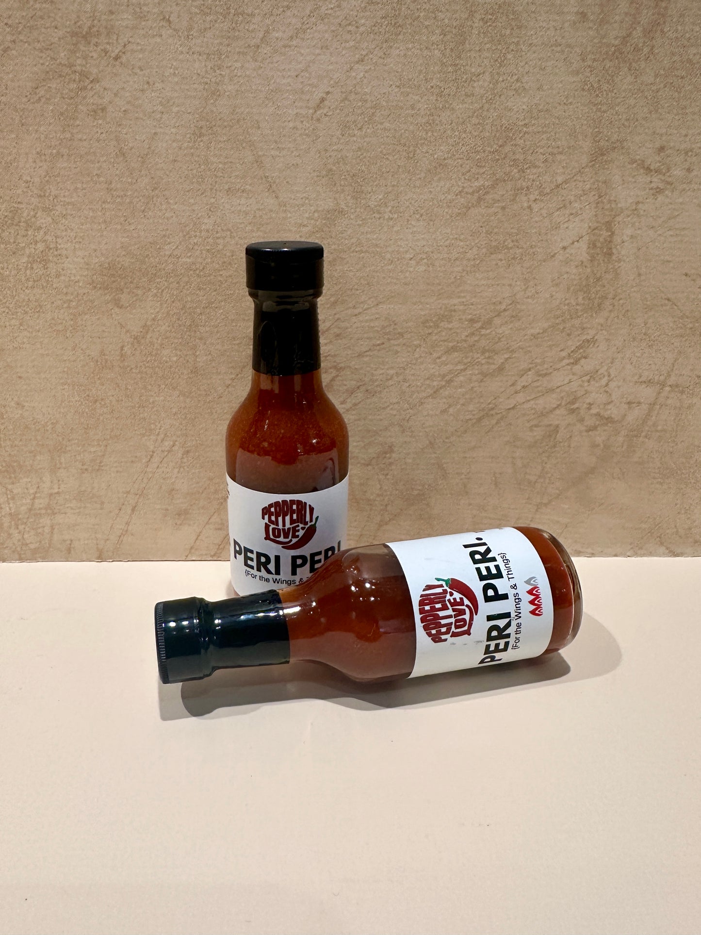 Two bottles of Peri Peri sauce on a beige surface with a wooden background
