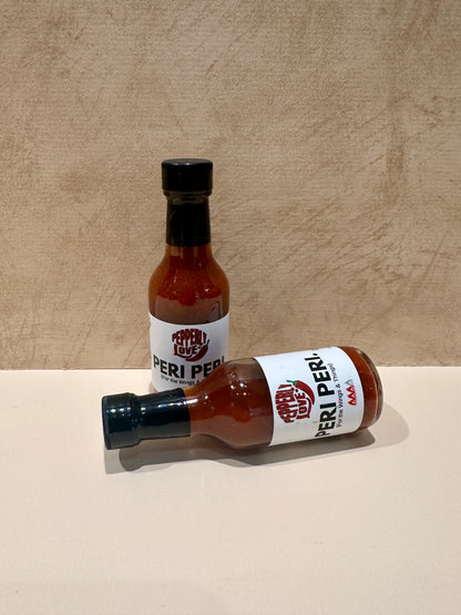 Two bottles of Peri Peri sauce on a beige surface with a wooden background