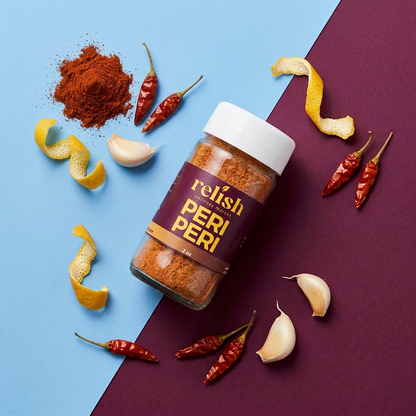 A bottle of Relish Market Peri Peri seasoning is displayed on a split blue and dark purple background, surrounded by raw ingredients including red chili peppers, garlic cloves, and lemon zest.