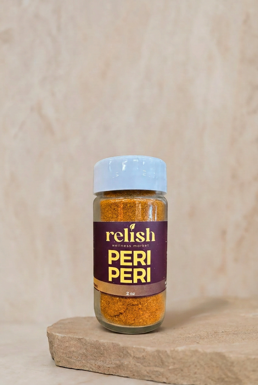 A bottle of Relish Market Peri Peri seasoning stands on a stone slab against a textured beige background. The bottle contains orange-red ground pepper, has a dark purple label with gold lettering, and a white cap.