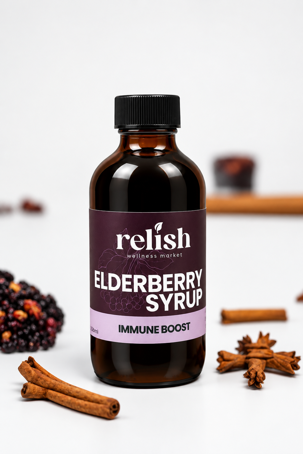 A bottle of Relish Market Elderberry Syrup labeled 'Immune Boost' stands on a white surface with cinnamon sticks, star anise, and dark berries arranged around it.
