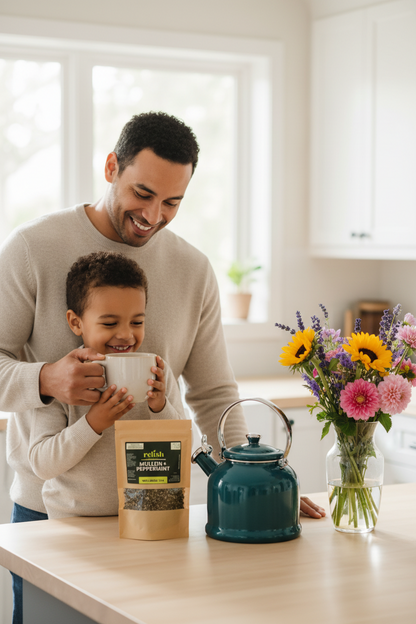 A man and a child smile while holding mugs in a bright kitchen, with a pouch of tea on the counter in front of them and a teal kettle and flower vase nearby.