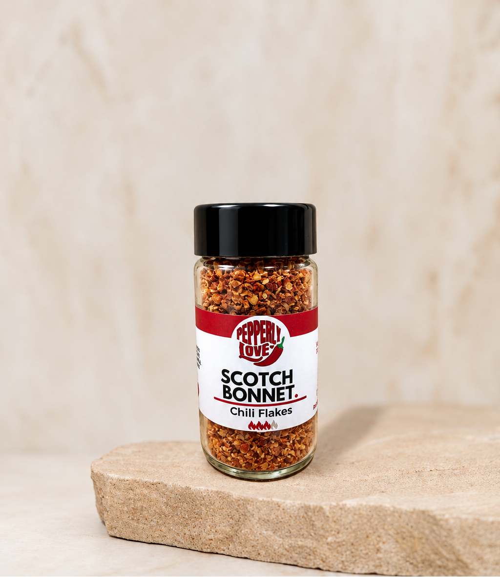 A small glass jar of Scotch Bonnet chili flakes by Pepperly Love with a black lid and white/red label, standing on a stone slab against a textured beige background.