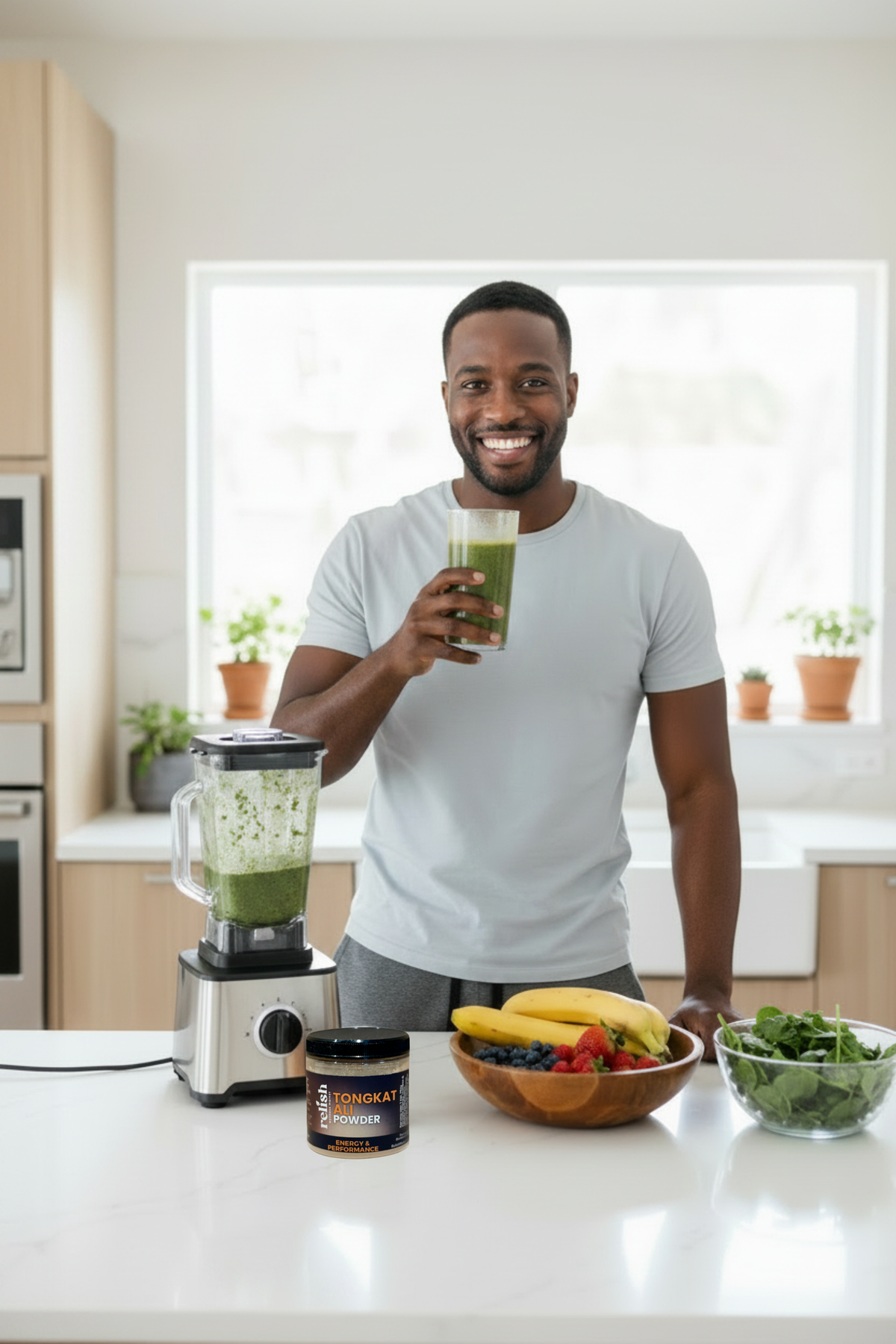 A smiling man in a white t-shirt stands in a bright kitchen holding a glass of green smoothie. On the counter in front of him is a green smoothie blender, a jar of Tongkat Ali Powder by Relish Market, a bowl of fruit, and a bowl of greens.